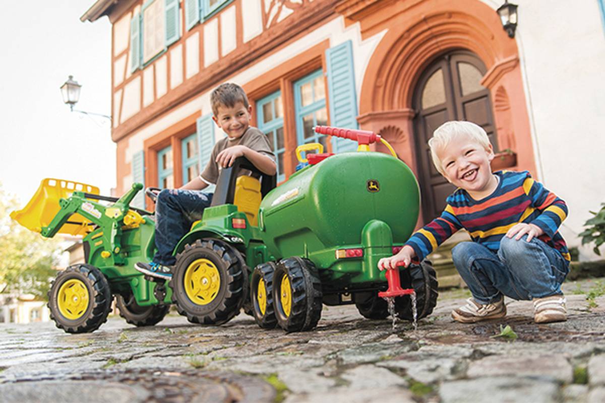 Zwei Kinder spielen draußen; eines fährt einen grünen Spielzeugtraktor, während das andere lachend an einem Spielzeugschlauch dreht. Im Hintergrund steht ein malerisches Haus.