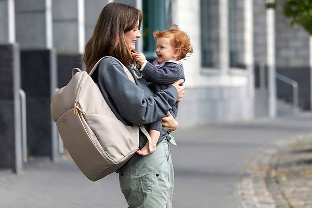 Frau hält im Freien ein lächelndes Baby und trägt einen großen beigen Rucksack. Beide wirken glücklich und stehen auf einem gepflasterten Weg mit Gebäuden im Hintergrund.