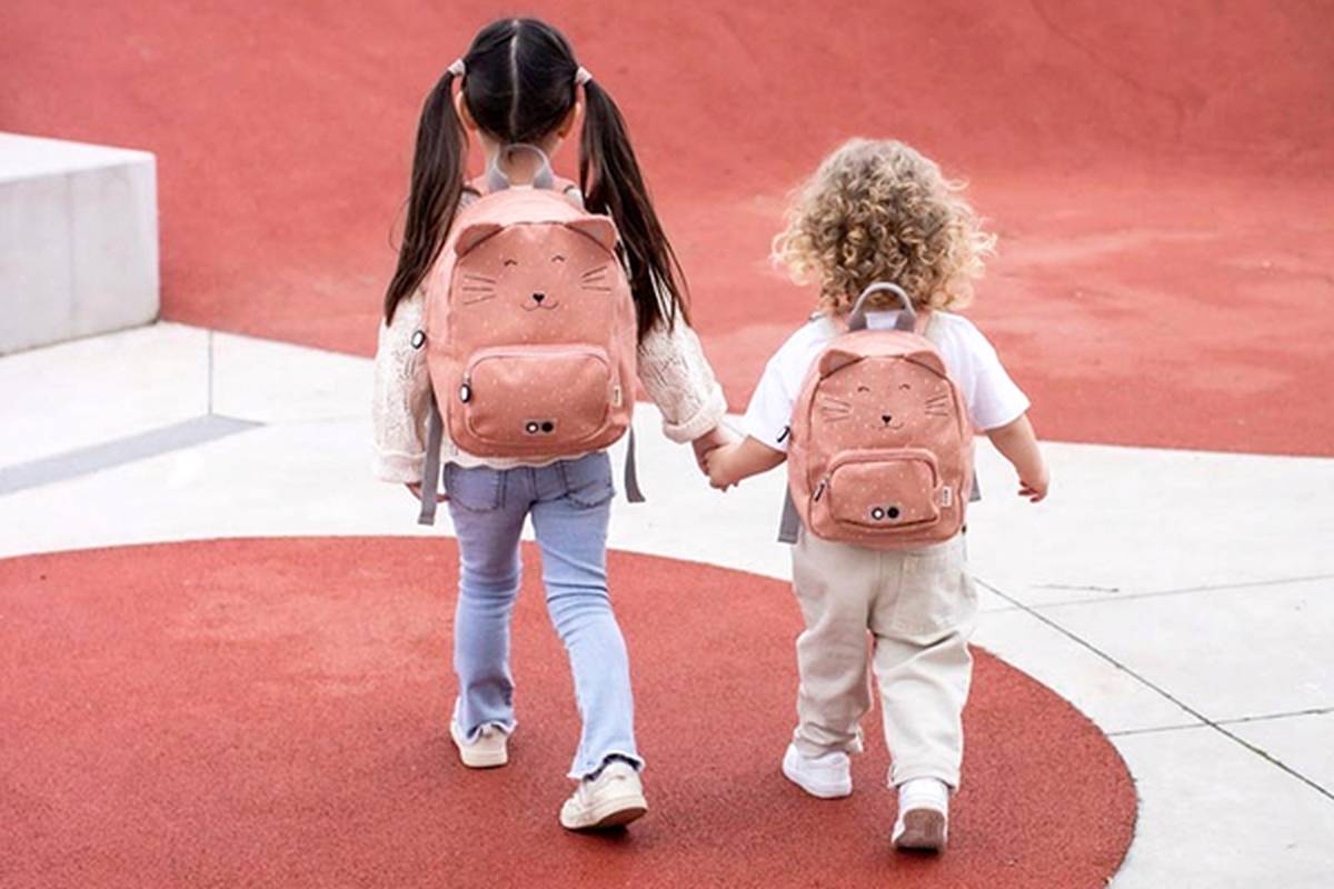 Zwei kleine Kinder mit Zöpfen und lockigem Haar, die Rucksäcke mit Tiermotiven tragen, halten Händchen, während sie auf einem rot-weißen Weg entlanggehen.
