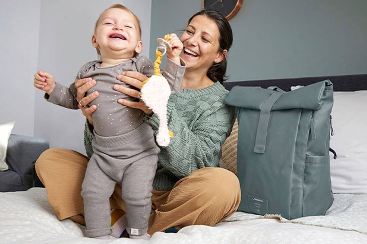Eine lächelnde Frau sitzt mit einem glücklichen Baby, das ein Spielzeug hält, auf dem Bett; daneben steht ein türkisfarbener Rucksack.