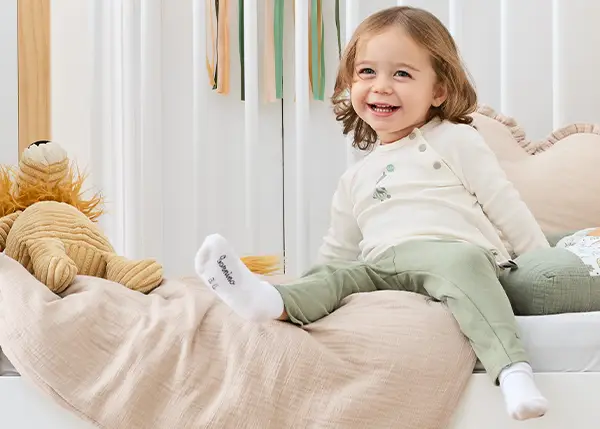 Ein lächelndes Kleinkind in einem weißen Oberteil und einer grünen Hose sitzt auf einem Bett mit einer beigen Decke und einem Spielzeuglöwen in der Nähe; im Hintergrund ist eine bunte Dekoration zu sehen.