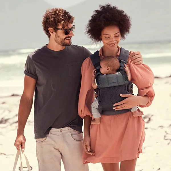 Ein Paar geht mit einem Baby in einer Trage am Strand spazieren. Die Frau trägt ein pfirsichfarbenes Kleid und der Mann eine Sonnenbrille sowie ein graues Hemd; sie lächeln sich gegenseitig an.