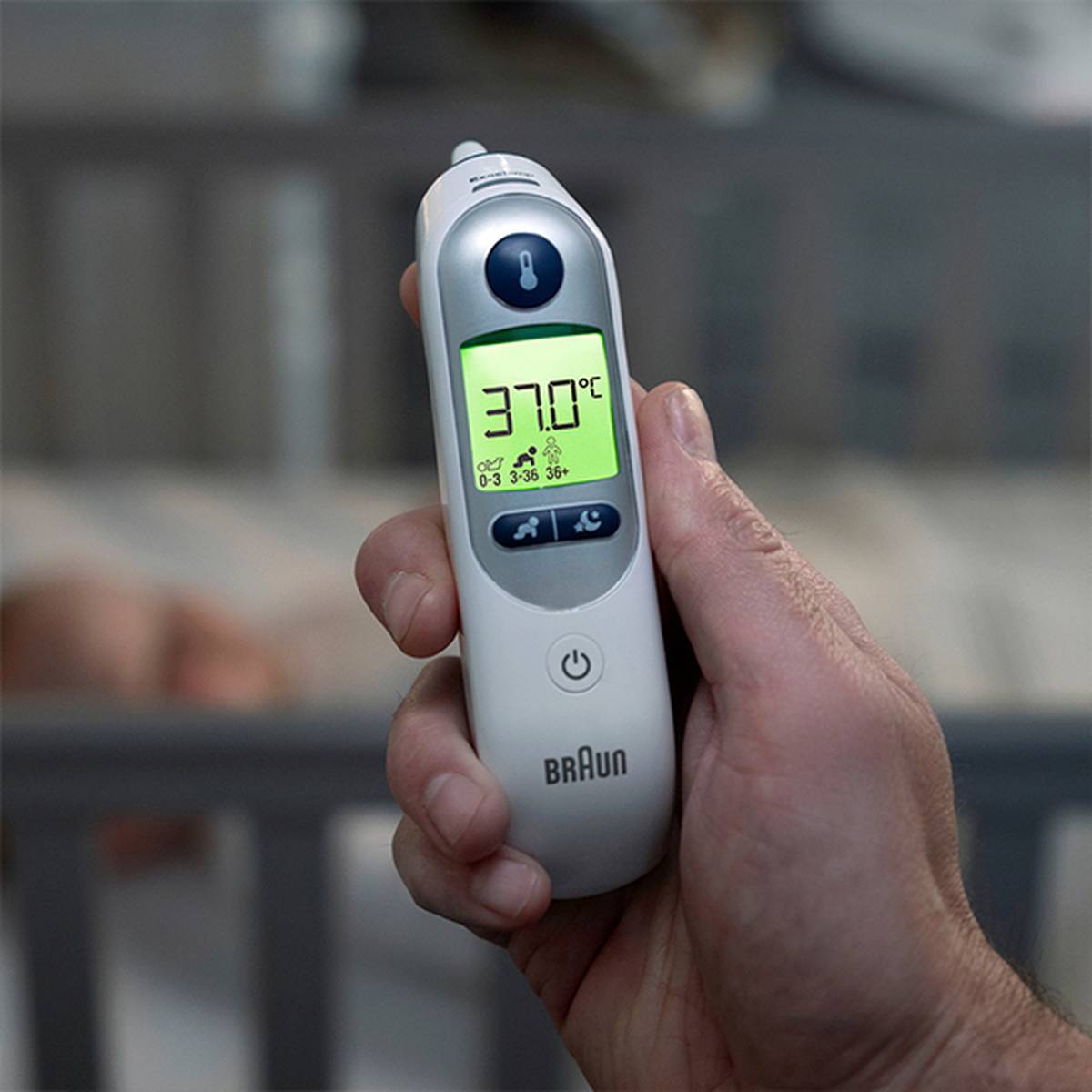 Nahaufnahme einer Hand, die ein digitales Braun-Thermometer hält, das 37,0 °C anzeigt, mit einem unscharfen Babybett im Hintergrund.