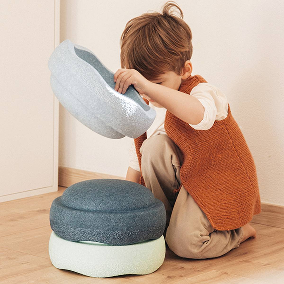 Ein Kind, das in einem Zimmer auf einem Holzboden sitzt und eine braune Weste und beige Hosen trägt, stapelt Stapelsteine.