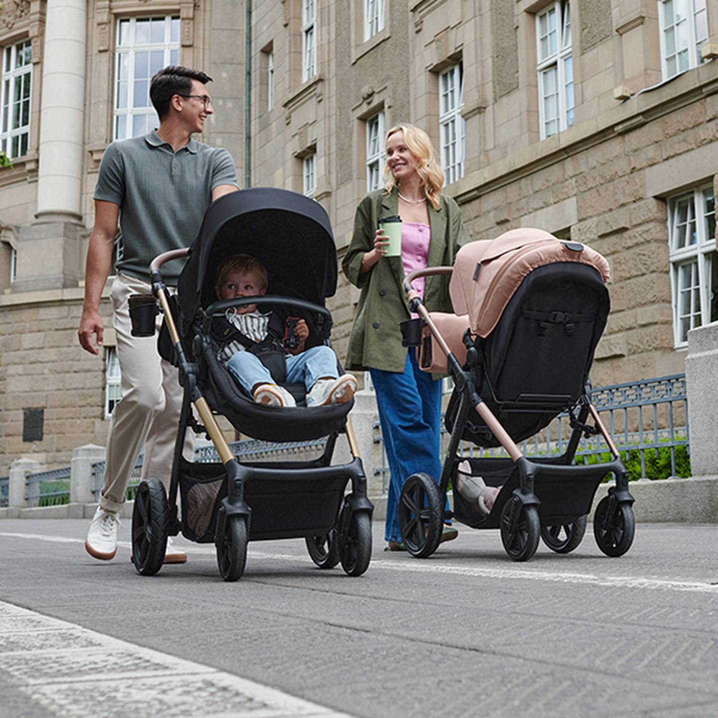 Ein Paar geht auf einer Stadtstraße spazieren. Jeder schiebt einen Kinderwagen mit einem Kind darin. Sie lächeln und genießen einen schönen Tag.