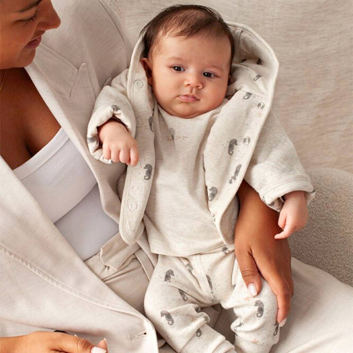 Eine Frau hält ein Baby im Arm, das ein gemütliches, hellfarbiges Outfit mit Mustern trägt und auf einem beigen Stuhl sitzt.