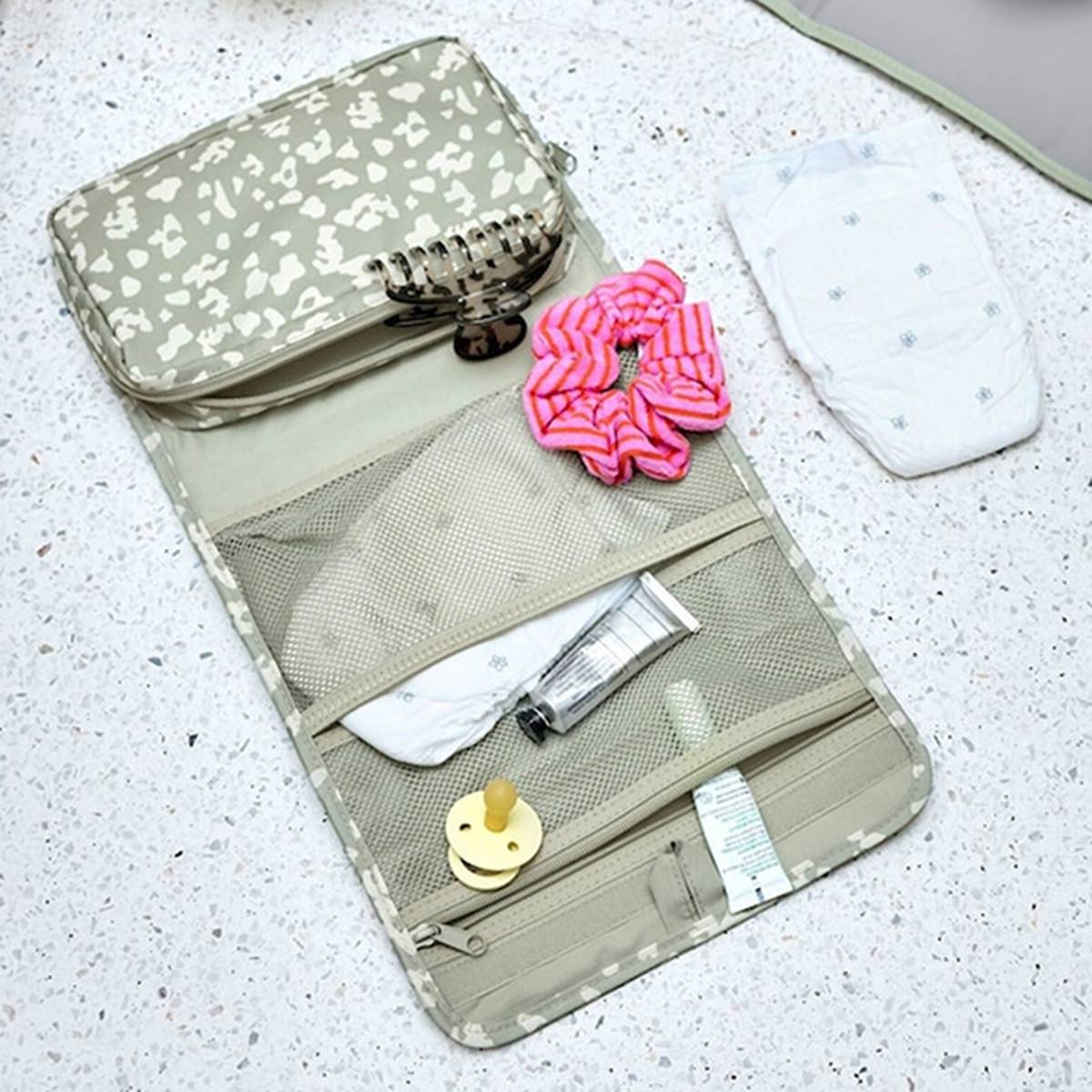 Offene Wickeltasche mit Fächern, in denen sich eine Haarspange, ein Haargummi, Creme, ein Schnuller und eine separate Windel befinden, auf einem gesprenkelten Boden.
