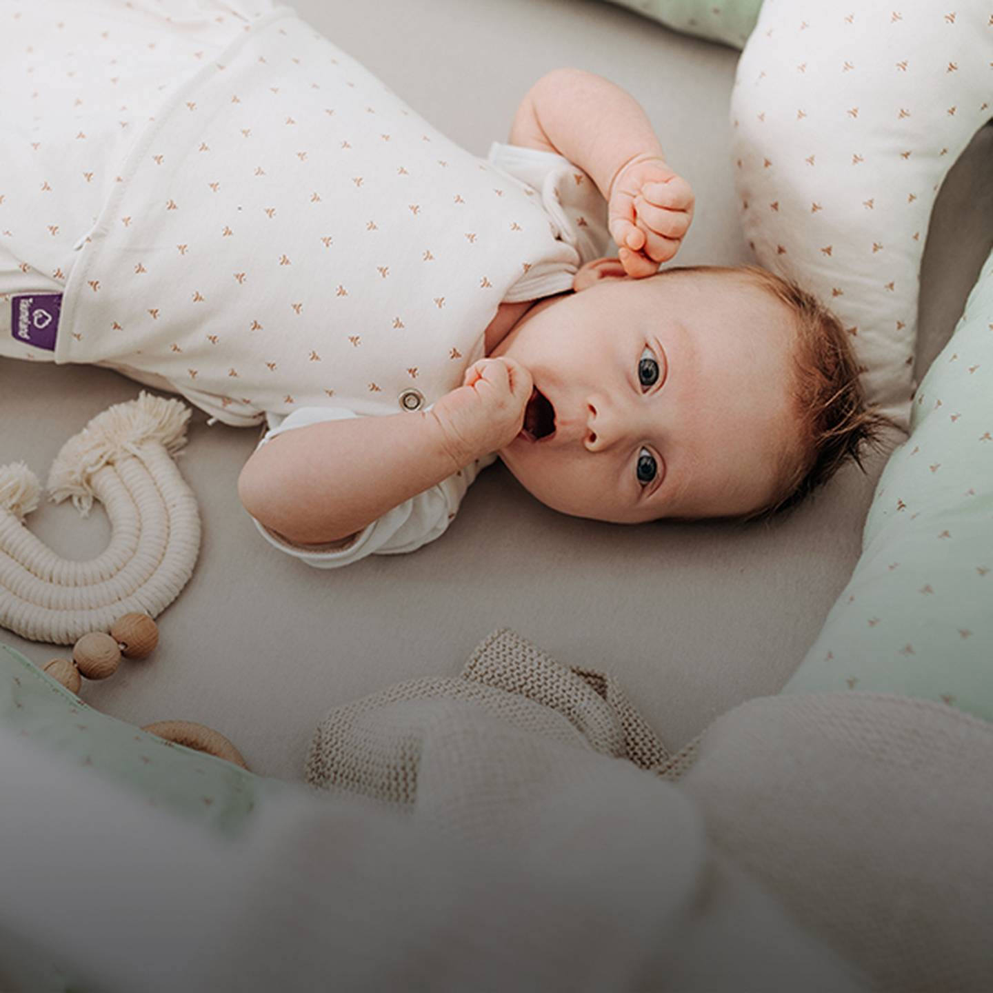 Baby liegt in einem Kinderbett, trägt einen weißen Strampler mit kleinen Mustern und ist von weicher mintgrüner Bettwäsche und Spielzeug umgeben.