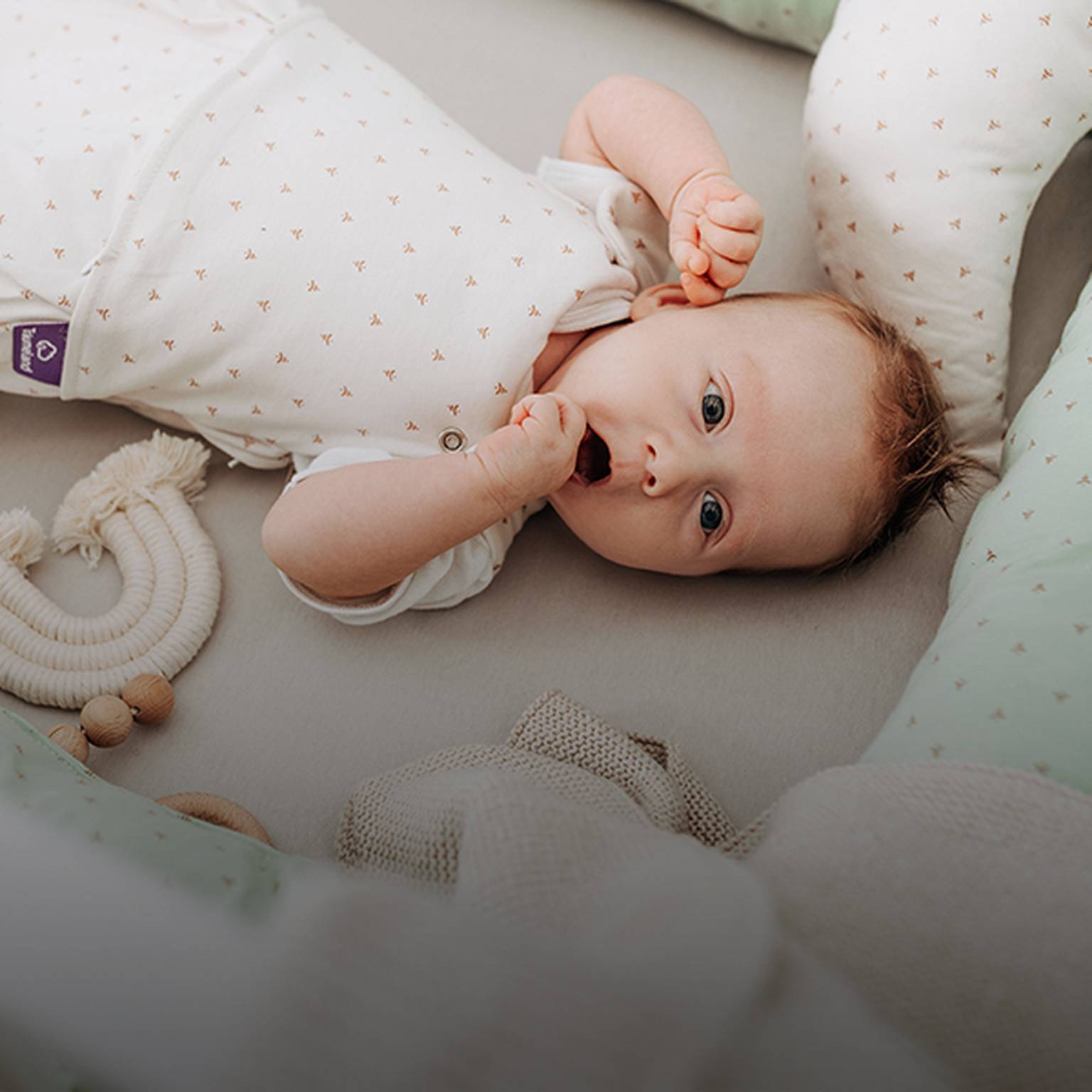 Baby liegt in einem Kinderbett, trägt einen weißen Strampler mit kleinen Mustern und ist von weicher mintgrüner Bettwäsche und Spielzeug umgeben.