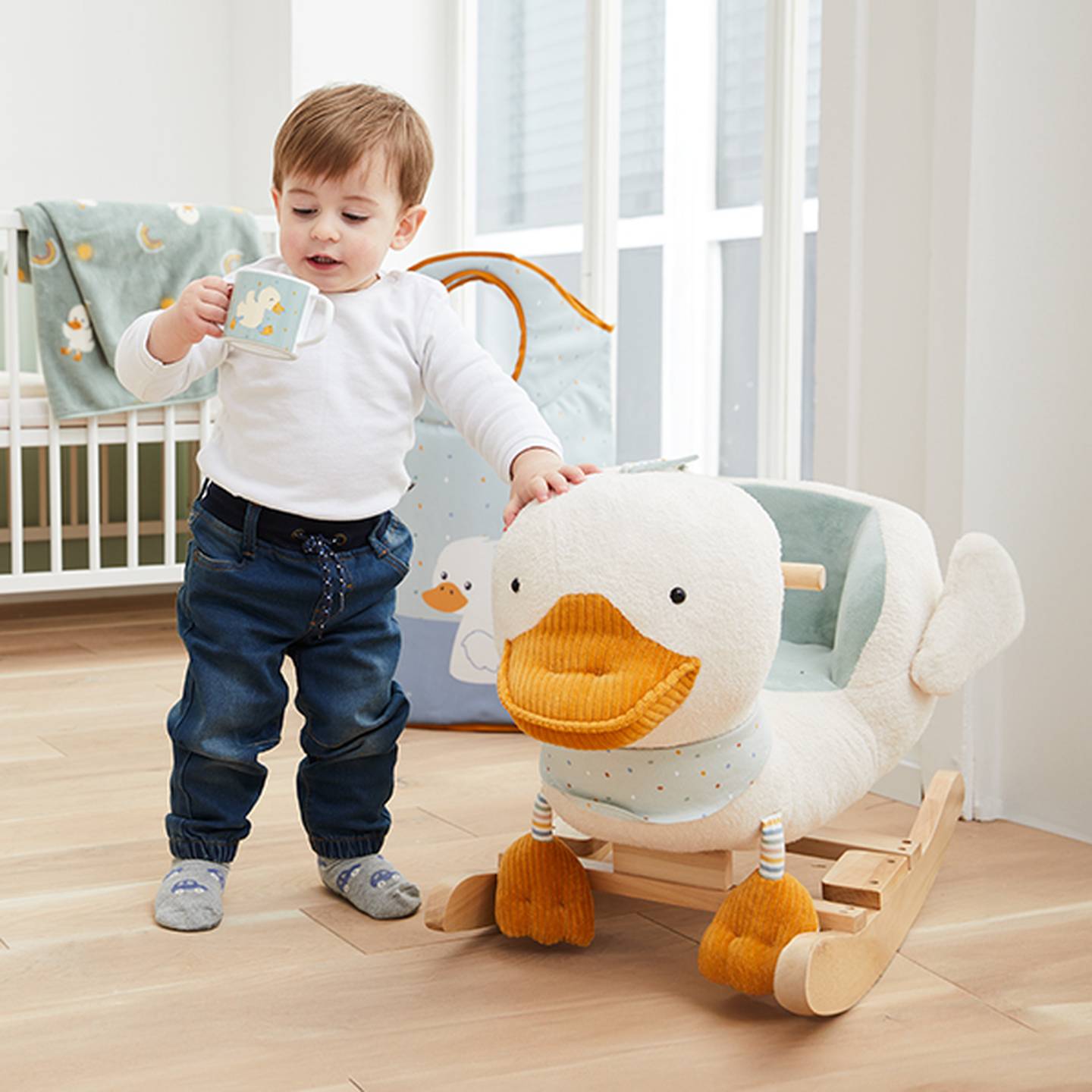 Ein Kleinkind mit einer Tasse steht neben einem Schaukelspielzeug in Entenform in einem Kinderzimmer mit Kinderbett und weicher Einrichtung.