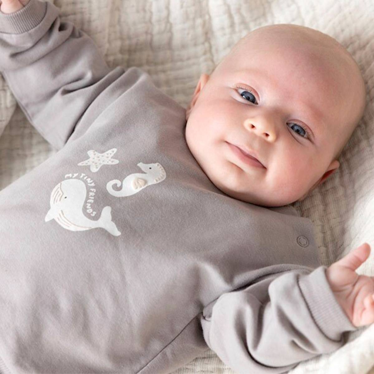 Ein Baby liegt auf einer weichen Decke und trägt einen grauen Strampler mit Meeresmotiven. Es blickt mit einem leichten Lächeln in die Kamera.
