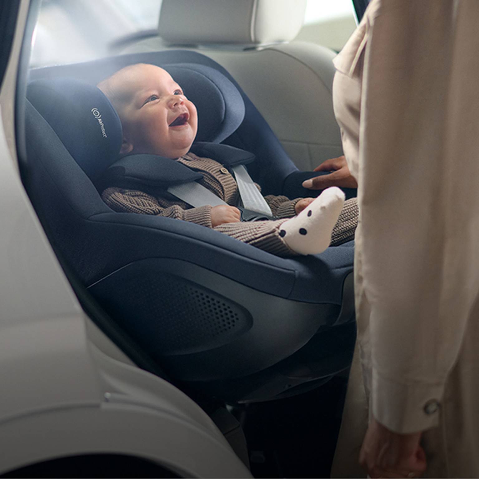 Ein lächelndes Baby in einem Autositz blickt zu einem Erwachsenen in einem Auto auf.