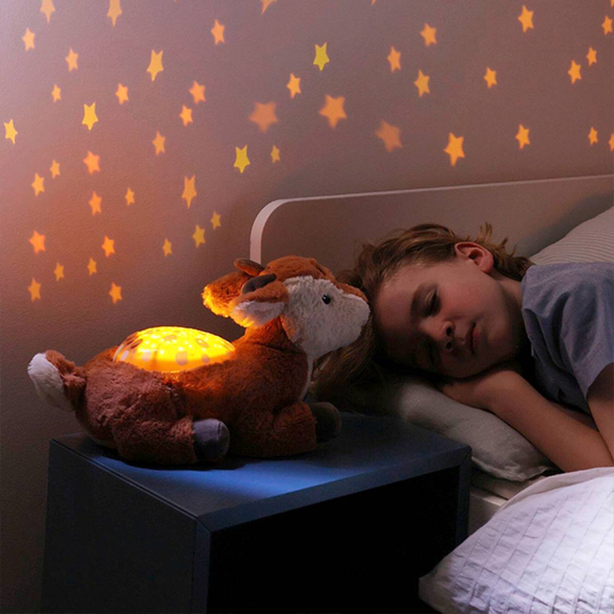 Kind schläft neben einem plüschigen Nachtlicht in Form eines Hirsches, das in einem schwach beleuchteten Raum leuchtende Sterne an die Wand und Decke projiziert.