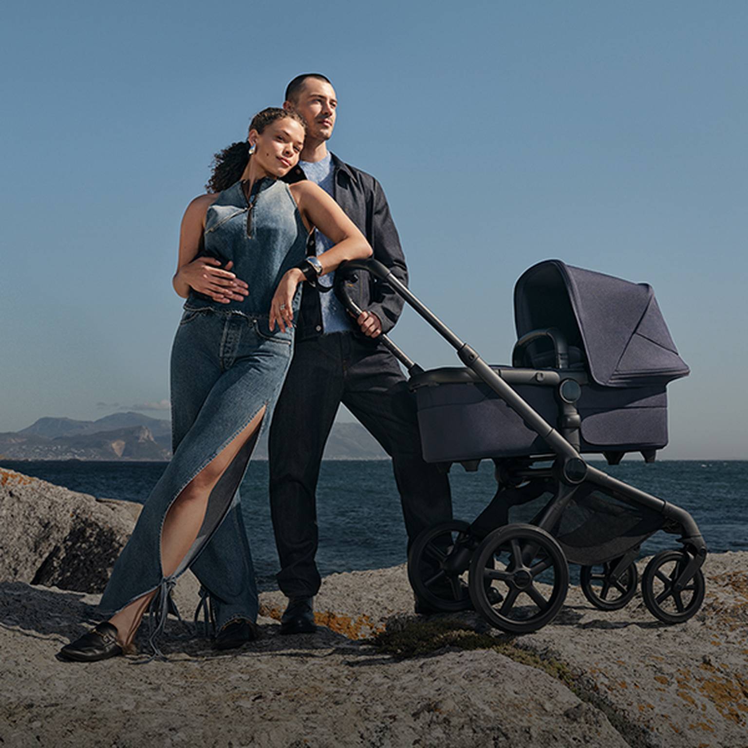 Paar in Jeanskleidung steht am felsigen Ufer mit einem marineblauen Kinderwagen, Meer und Berge im Hintergrund unter einem klaren blauen Himmel.