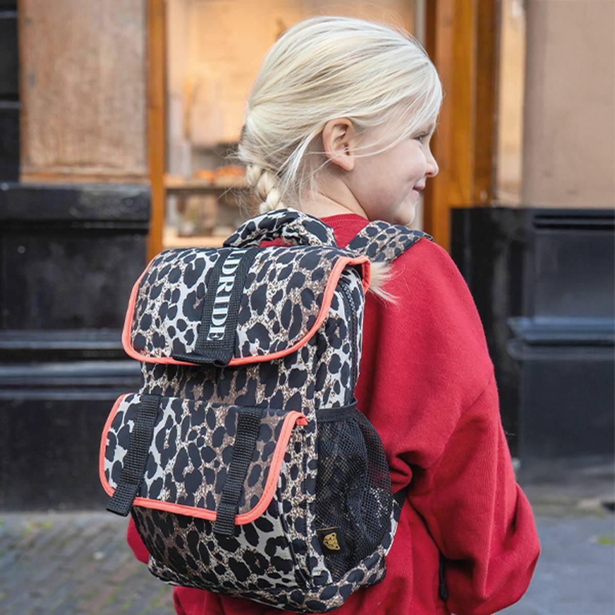 Lächelndes Kind, das einen roten Kapuzenpulli und einen Rucksack mit Leopardenmuster trägt und vor einem Gebäude mit großen Fenstern steht.