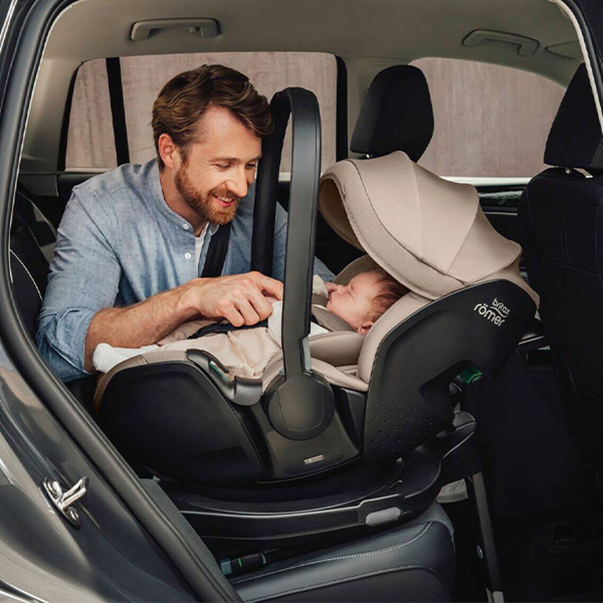 Ein Mann lächelt einem schlafenden Baby in einem Autositz zu und verstellt den Griff. Der Autositz ist im Fahrzeug nach hinten gerichtet.