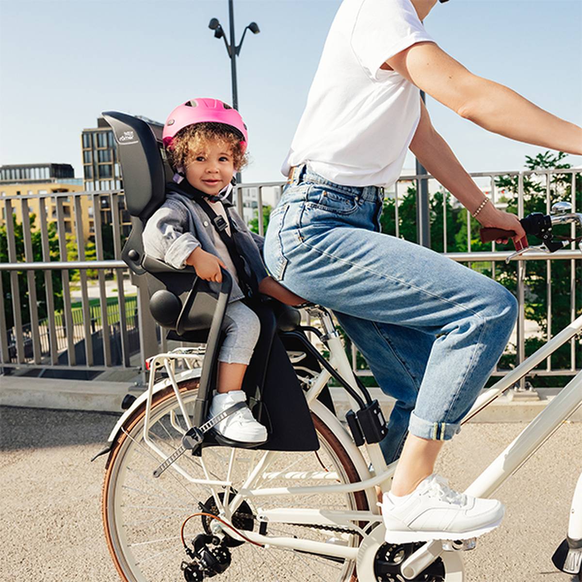 Erwachsener mit Kind im Kindersitz auf dem Fahrrad. Beide tragen Helme; das Kind einen rosa, der Erwachsene einen weißen. Im Hintergrund sind städtische Gebäude und Bäume zu sehen.