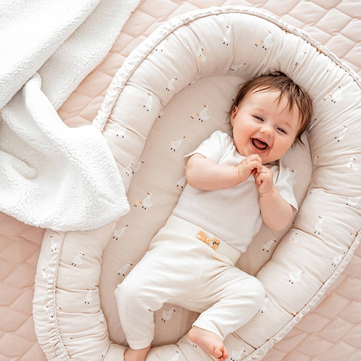 Ein lächelndes Baby in einem weißen Outfit liegt in einem weichen, gemusterten Babynest, neben ihm eine weiße Decke.