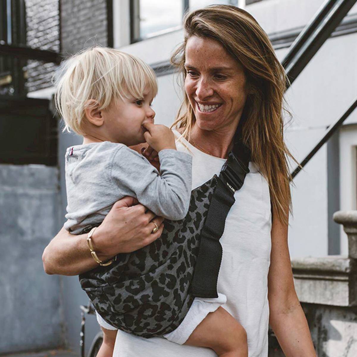 Frau lächelt, während sie ein kleines Kind in einem Tragetuch mit Leopardenmuster im Freien trägt. Das Kind hat blonde Haare und einen Finger im Mund.
