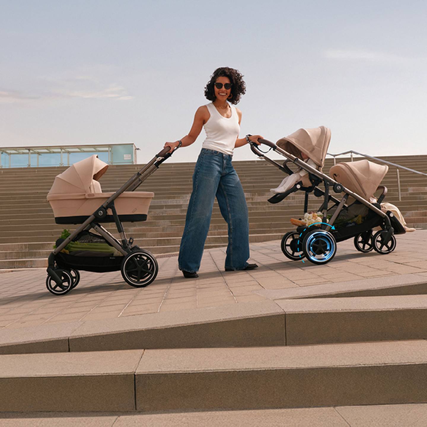Eine Frau in einem weißen Tanktop und Jeans schiebt zwei beige Kinderwagen auf einem gepflasterten Platz; im Hintergrund sind Treppen zu sehen.