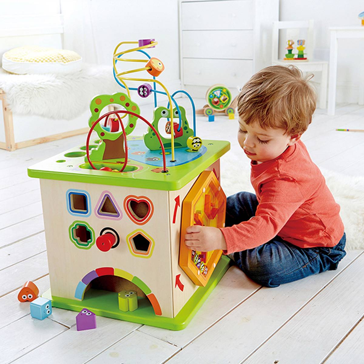 Ein Kind spielt auf einem weißen Holzboden mit einem bunten Aktivitätswürfel, der Formensortierspiele, Perlenlabyrinthe und interaktive Elemente enthält.