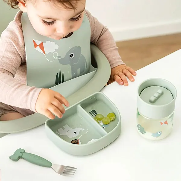 Ein Kleinkind mit einem Lätzchen mit Elefantenmotiv sitzt in einem Hochstuhl und greift nach Essen auf einem unterteilten Tablett; daneben steht ein Trinkbecher.