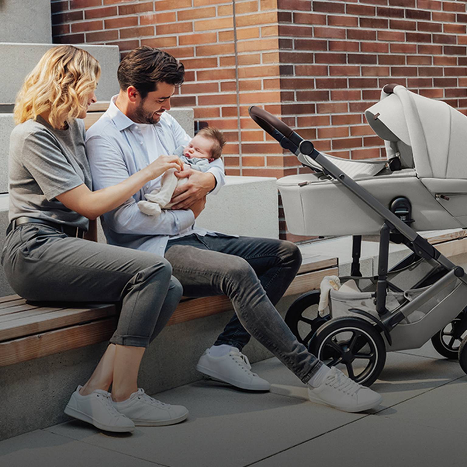 Ein Paar sitzt auf einer Bank und lächelt sein Baby an. Der Vater hält das Baby im Arm, daneben steht ein Kinderwagen. Es ist ein sonniger Tag.