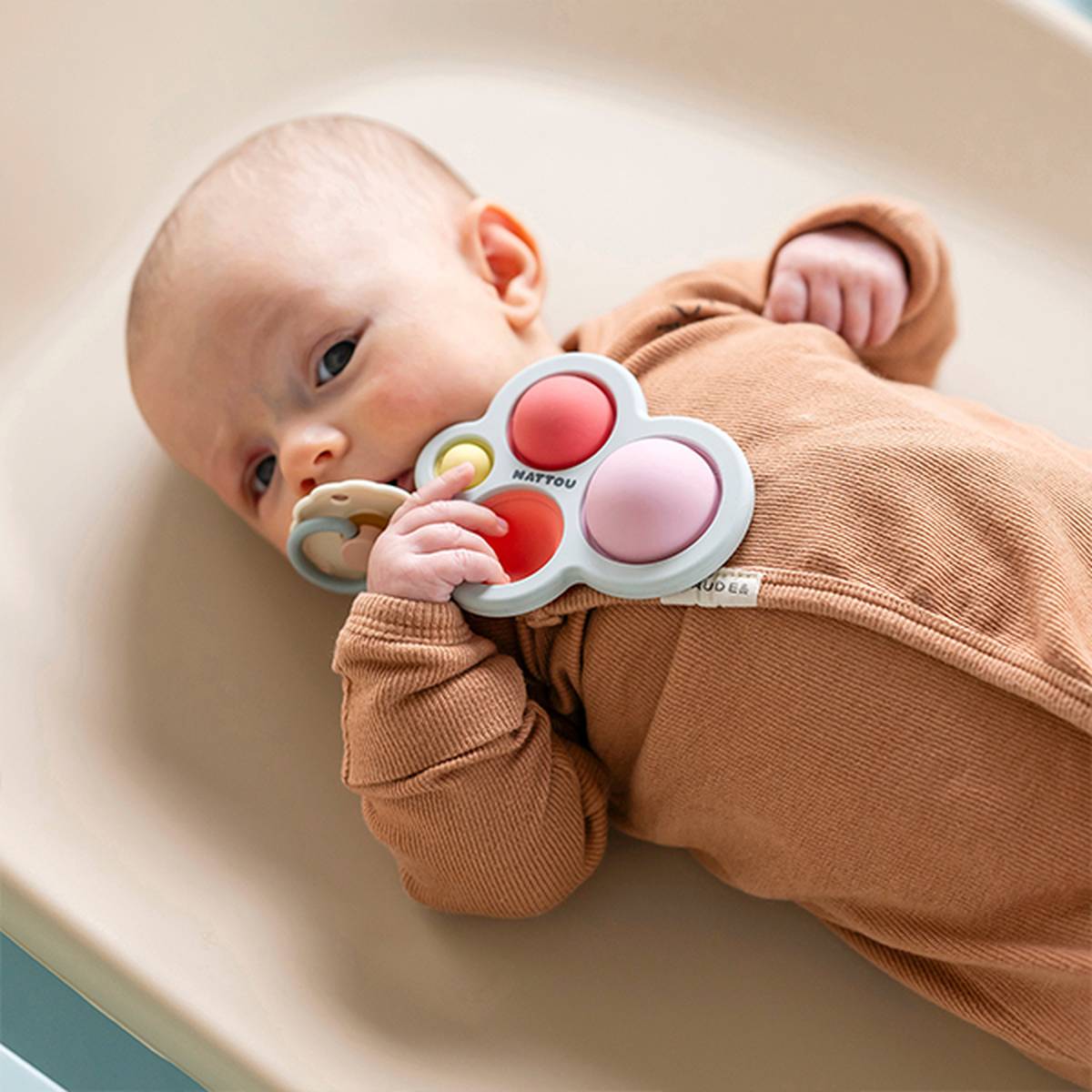 Ein Baby in einem braunen Strampler hält ein buntes Beißspielzeug, während es auf einer beigen Unterlage liegt.