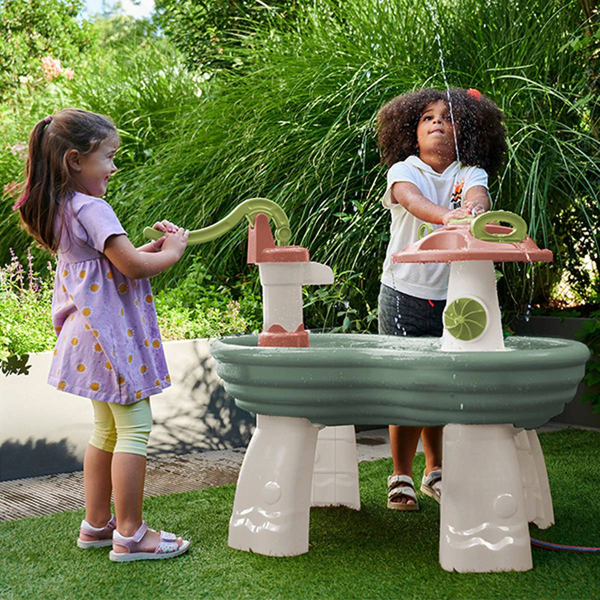 Zwei Kinder spielen an einem Wassertisch in einem Garten, eines pumpt Wasser, das andere fängt es auf, umgeben von grünen Pflanzen.
