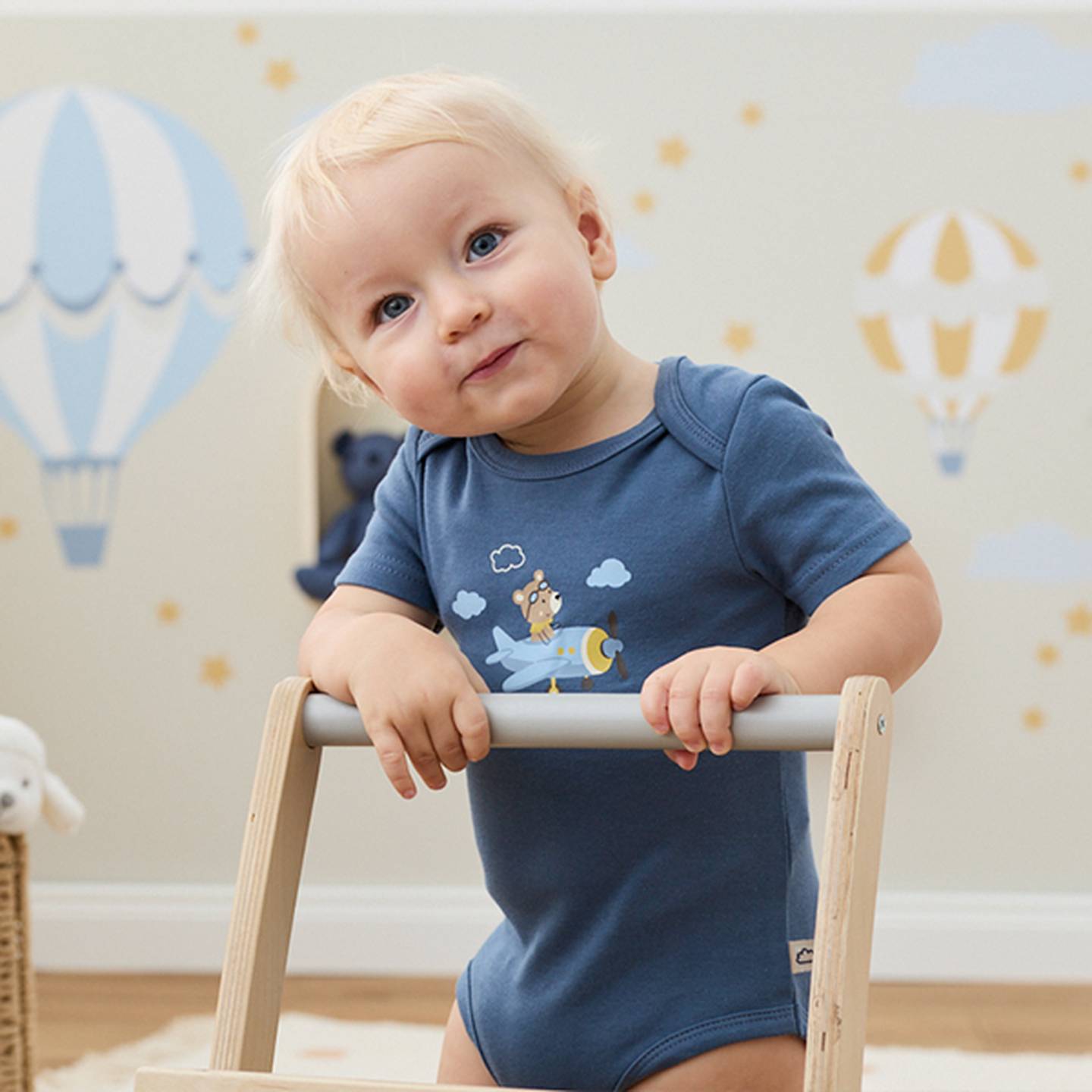 Ein Baby mit blonden Haaren lehnt sich an einen hölzernen Lauflernwagen, trägt einen blauen Strampler; im Hintergrund sind Wanddekorationen mit Heißluftballons zu sehen.
