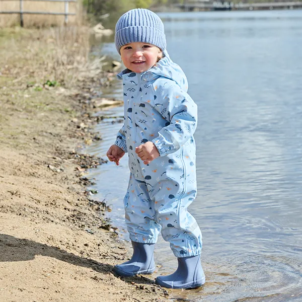 Ein Kind in blauer Kleidung und Hut steht am Wasserrand und trägt dazu passende Stiefel an einem sandigen Ufer.