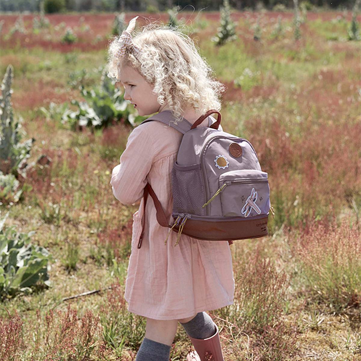 Junges Mädchen mit lockigem Haar, rosa Kleid und braunem Rucksack, geht durch eine Wiese.