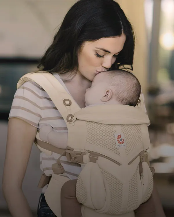 Eine Mutter küsst ihr Baby, das in einer beigen, ergonomischen Babytrage schläft. Die Szene strahlt Wärme und Zärtlichkeit aus.