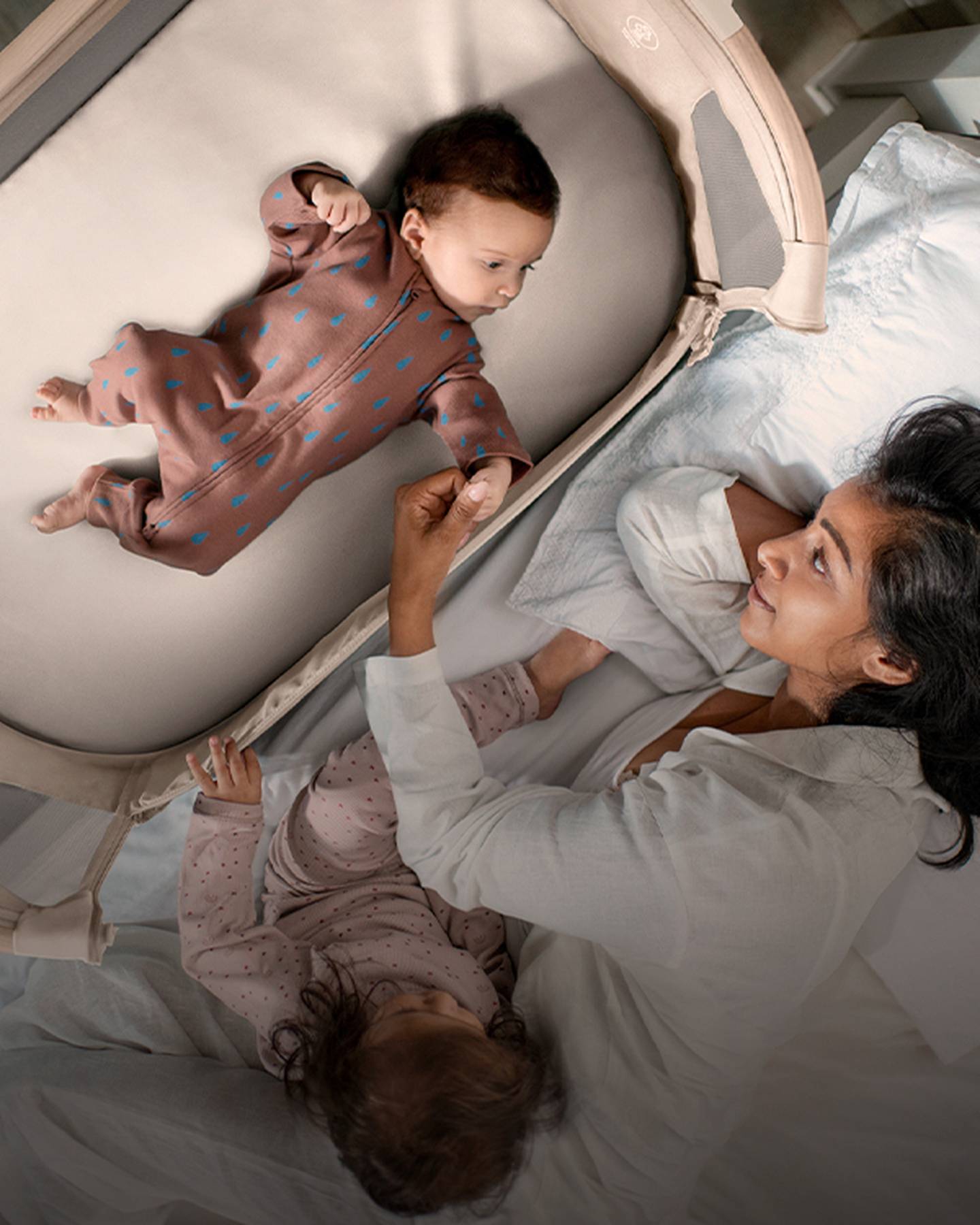 Eine Mutter liegt im Bett und hält ein Baby in der Wiege an der Hand. Das Baby trägt einen braunen Strampler mit blauen Punkten. Tageslicht durchflutet den Raum.