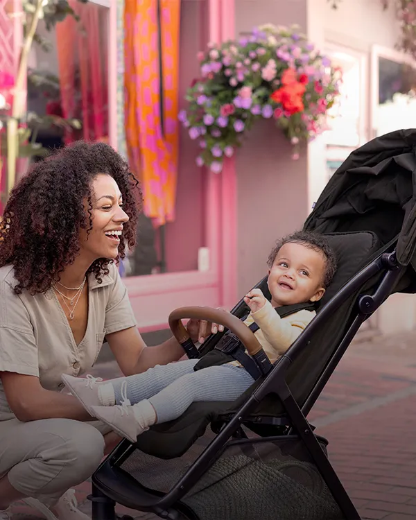 Eine Frau lächelt ein Baby in einem Kinderwagen auf einem Bürgersteig an; im Hintergrund sind rosafarbene Schaufenster und Pflanzen zu sehen.