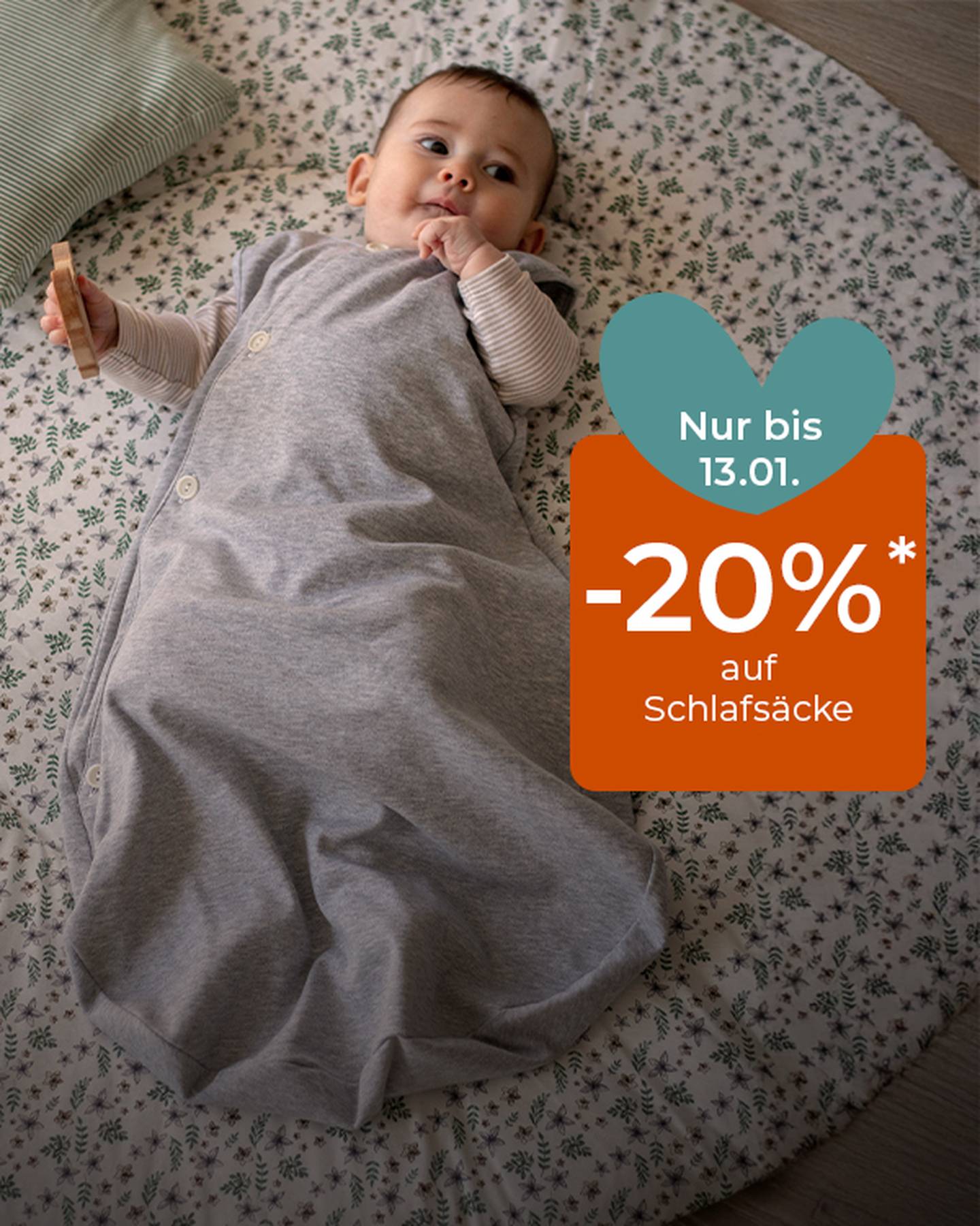 Baby liegt in einem grauen Schlafsack auf einer geblümten Matte und hält einen Beißring. Texteinblendung: „Nur bis 13.01. -20 %* auf Schlafsäcke.“
