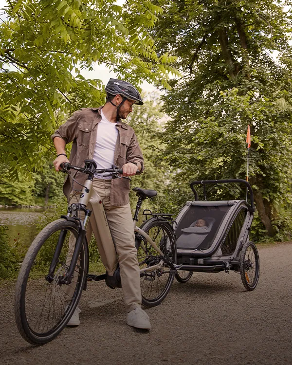 Ein Mann mit Fahrrad und Kinderanhänger steht auf einem von Bäumen gesäumten Weg und blickt zurück.