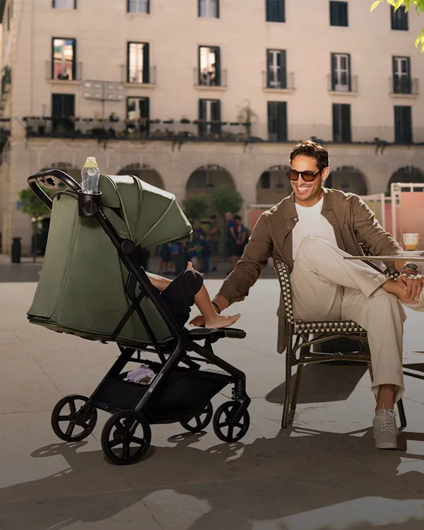 Ein Mann mit Sonnenbrille sitzt in einem Straßencafé und schiebt einen Kinderwagen mit einem Baby unter einem grünen Baldachin vor dem Hintergrund einer Stadtkulisse.