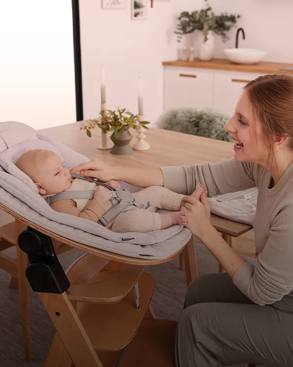 Eine Frau lächelt und interagiert mit einem Baby, das in einem gepolsterten Sitz auf einem Esstisch in einem gemütlichen Zimmer liegt.
