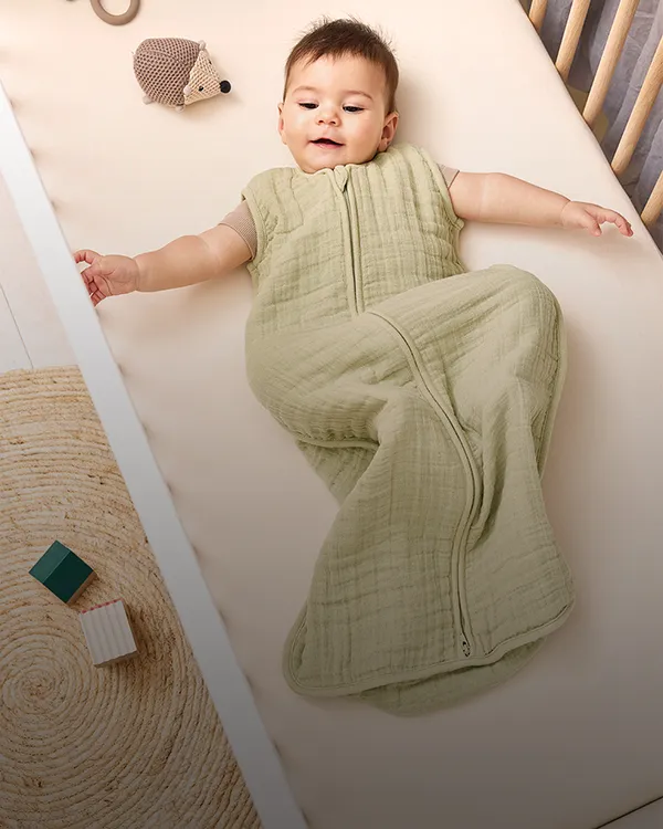 Baby im grünen Schlafsack liegt mit einem Teddybären und einem Beißring im Kinderbett und schaut nach oben.