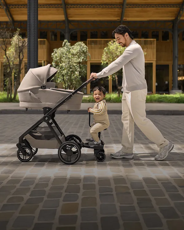Ein Mann schiebt einen Kinderwagen, auf dem ein Kleinkind auf einem Mitfahrbrett steht; beide lächeln. Die Straße ist mit Kopfsteinpflaster belegt, im Hintergrund sind Grünanlagen und Gebäude zu sehen.