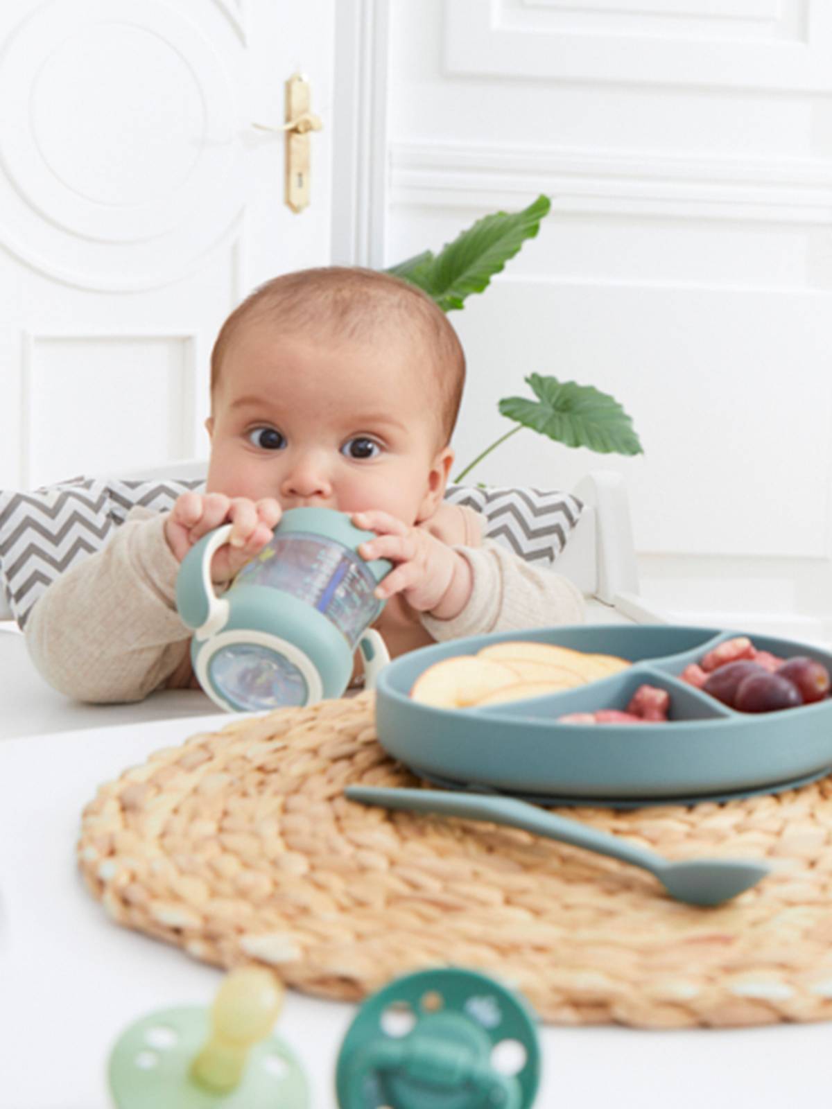 Baby sitzt an einem Tisch, hält einen Trinkbecher in der Hand, davor steht auf einem gewebten Tischset ein Teller mit Obst und Snacks.
