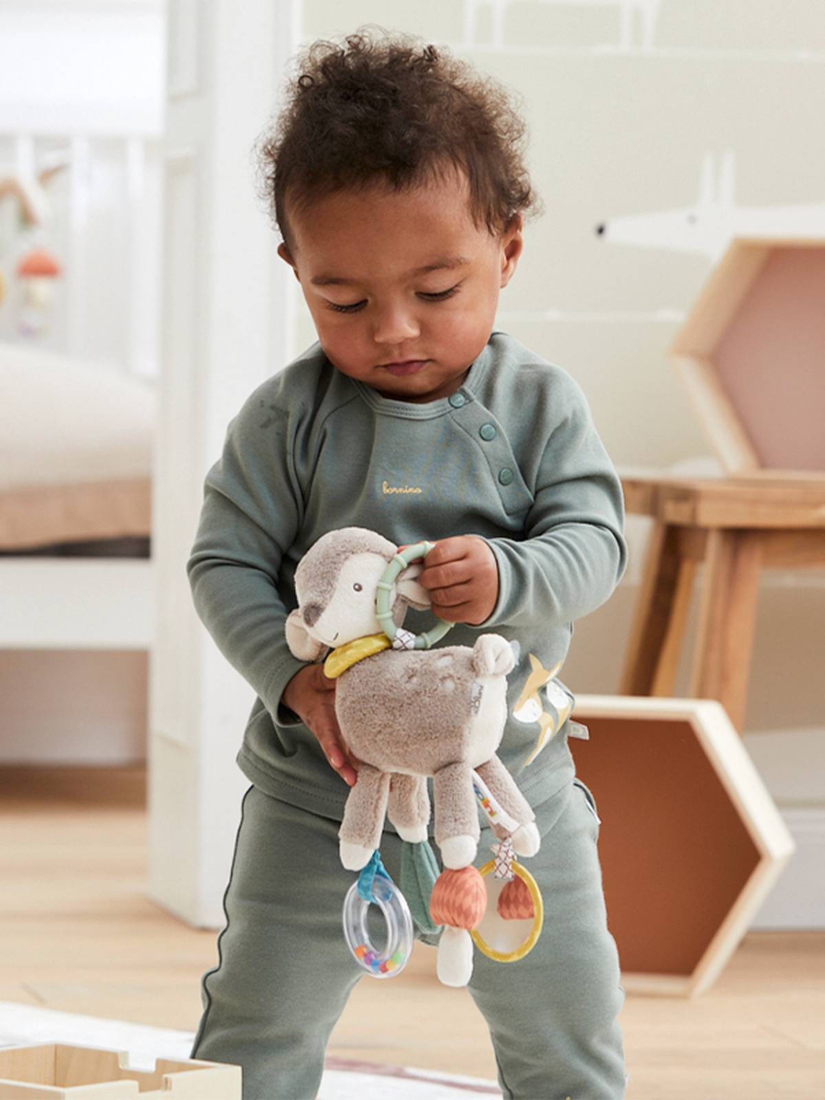 Ein Kleinkind in einem grünen Outfit hält ein Plüschtier mit Ringen und Rasseln und steht in einem gemütlichen Zimmer mit Holzmöbeln.