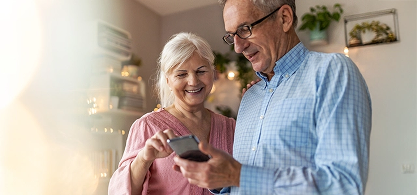 Zwei ältere Erwachsene stehen in einem Wohnraum und betrachten gemeinsam ein Smartphone, während Pflanzen und Regale im Hintergrund zu sehen sind.