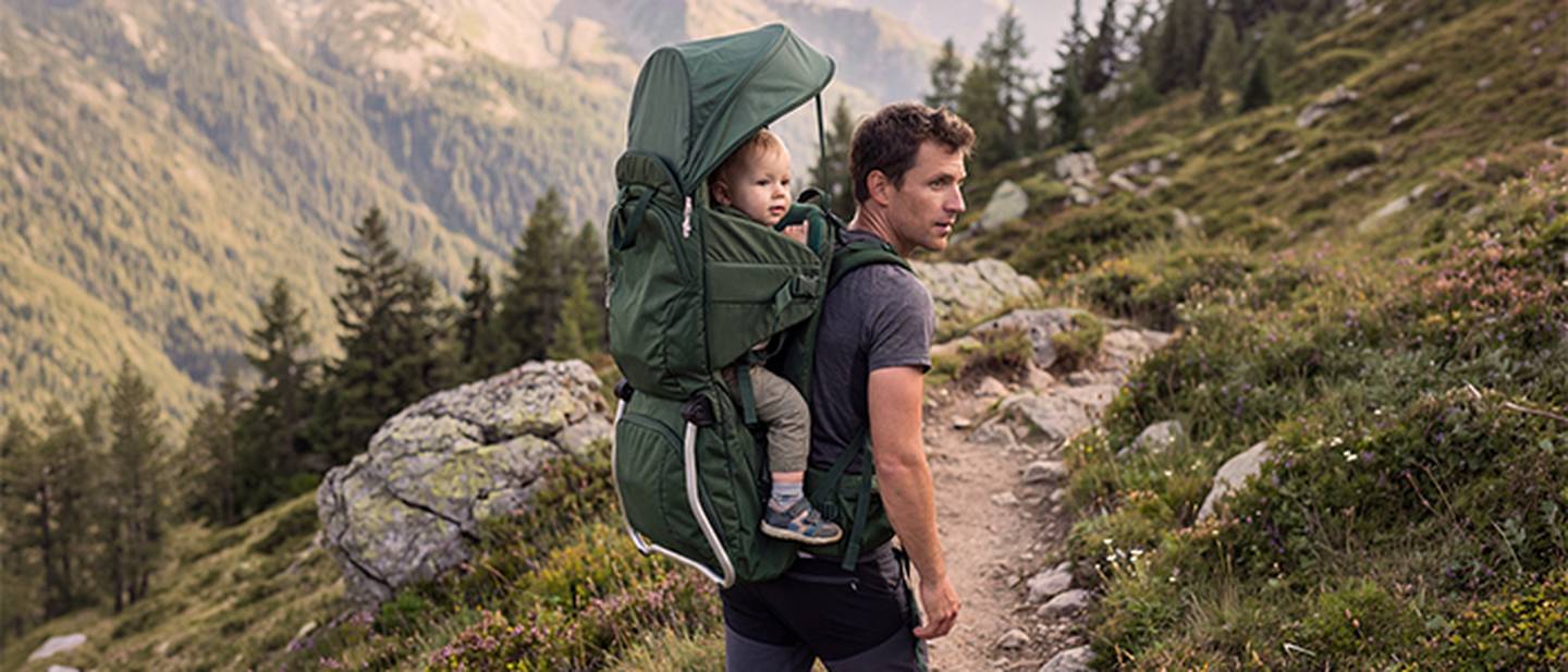 Ein Mann wandert auf einem Bergpfad und trägt ein Kleinkind in einer grünen Rückentrage. Üppiges Grün und ferne Gipfel umgeben sie.