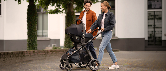 Ein Paar lächelt, während es draußen spazieren geht und einen Kinderwagen schiebt. Die Umgebung ist städtisch, mit Bäumen und Gebäuden im Hintergrund.