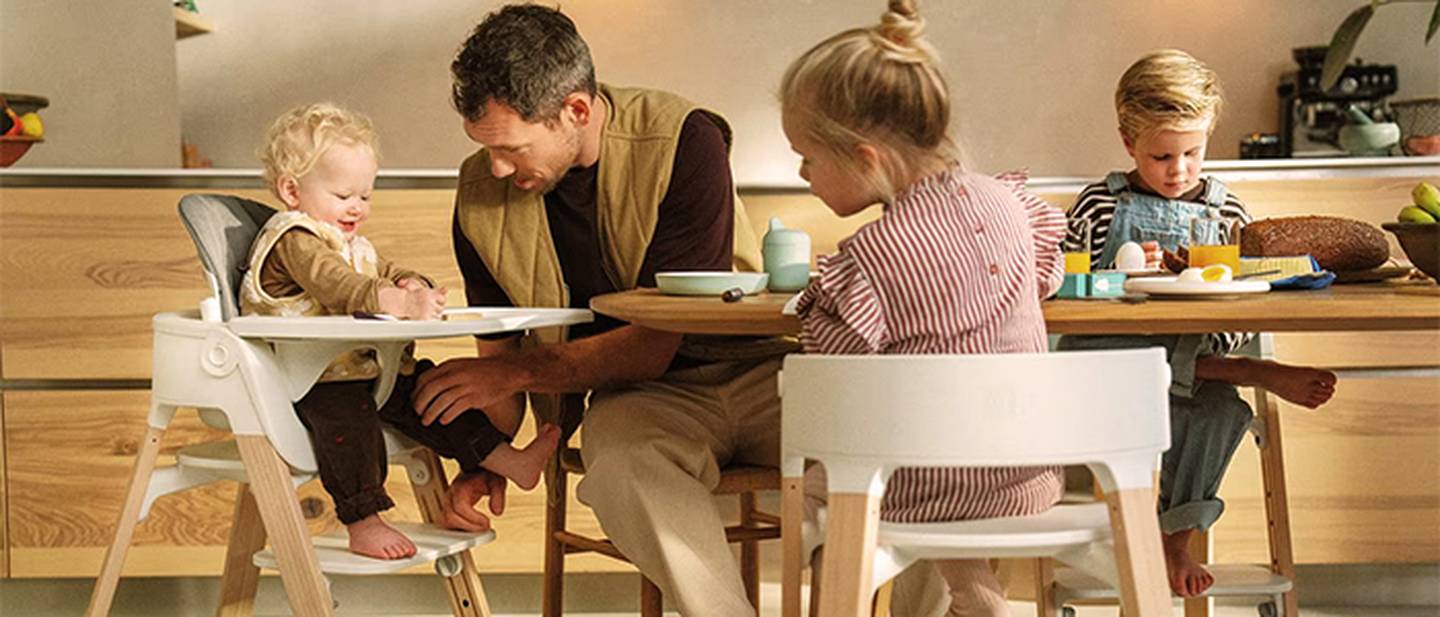 Eine Familie in der Küche, ein Mann und drei Kinder. Ein Kind sitzt in einem Hochstuhl, die anderen sitzen an einem Tisch mit Essen und Besteck.
