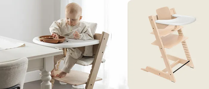 Ein Baby sitzt in einem hölzernen Hochstuhl mit einem weißen Tablett und isst von einem braunen Teller. Neben dem Baby steht ein leerer Hochstuhl.