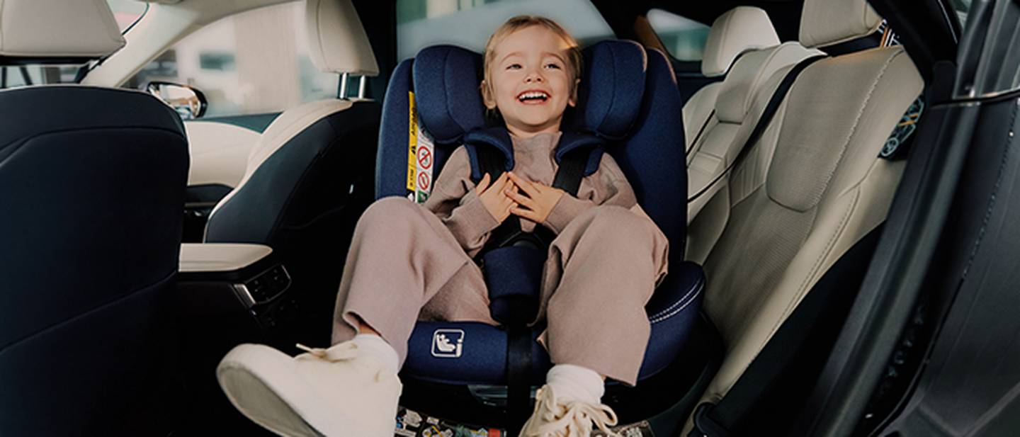 Ein lächelndes Kind sitzt sicher in einem blauen Kindersitz in einem Fahrzeug. Es trägt ein gemütliches Outfit und Turnschuhe.