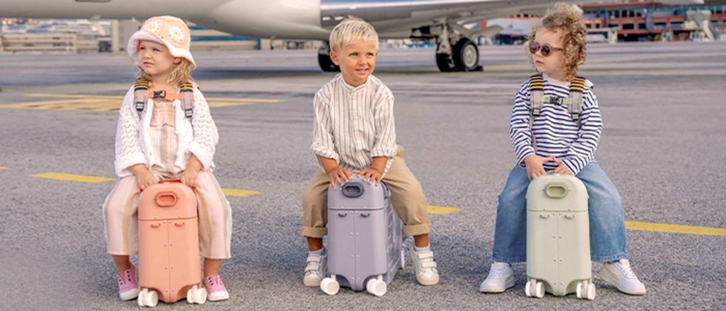 Drei Kinder sitzen auf bunten Koffern auf einer Flughafenlandebahn, im Hintergrund ist ein Flugzeug zu sehen.